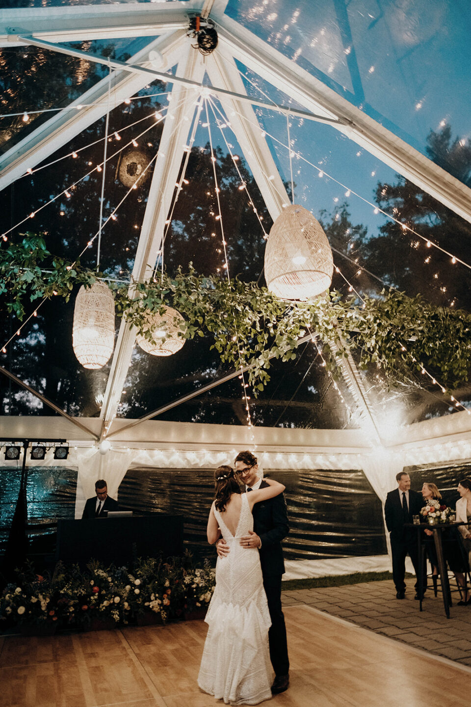 newlyweds have first dance under clear top tent with twinkle lights at night.