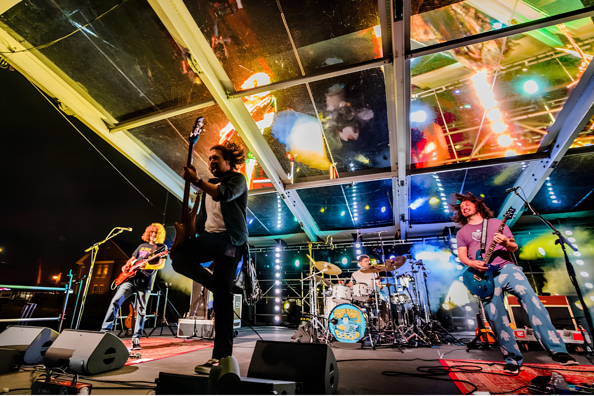 Live performance photo of the band Pigeons Playing Ping Pong performing on stage underneath a custom clear top vinyl stage roof skin manufactured by Select Event Group, showing stage lighting reflections through the transparent roof panels.