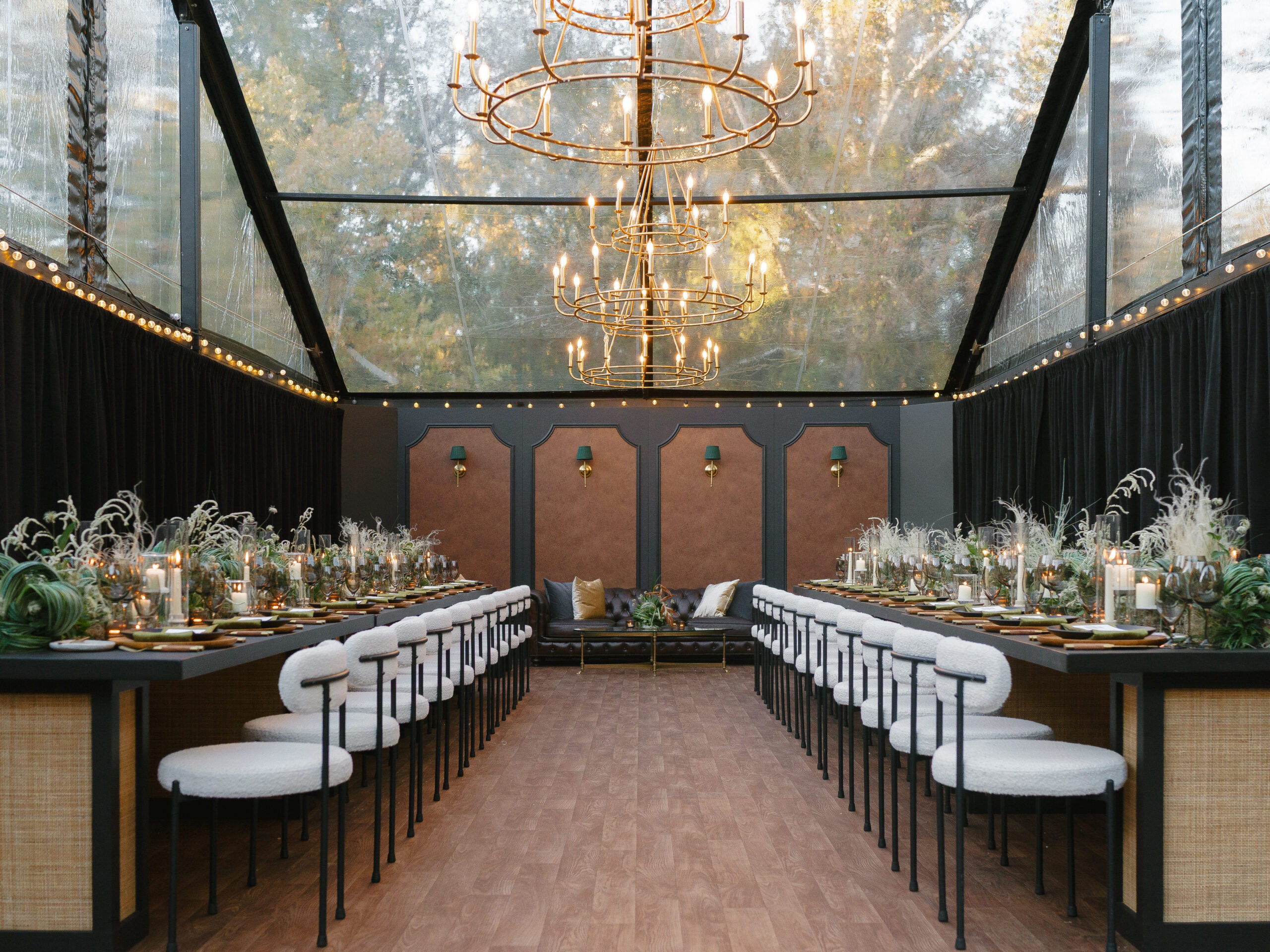 Clear-top tent set for a birthday dinner, with two long tables, white boucle chairs, gold candle chandeliers and a leather lounge sofa at the far end.