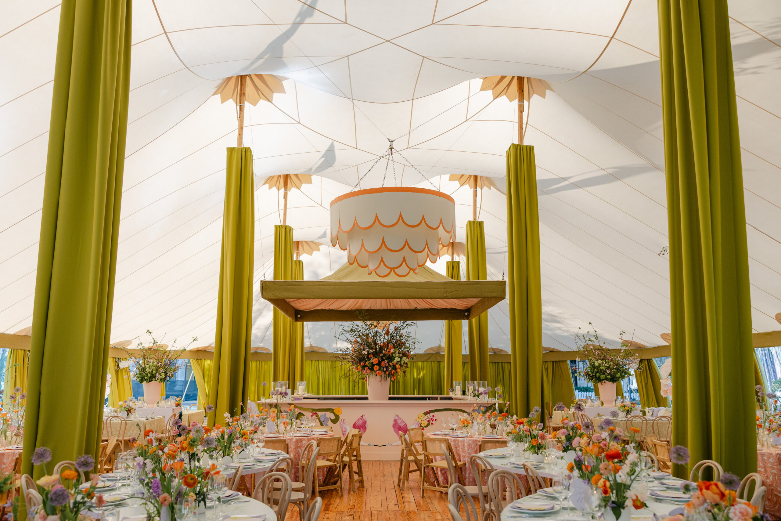 Sailcloth tent interior with round dinner tables, colorful flower centerpieces, green draped tent poles and a scalloped canopy over the bar. It shows how an event tent size fits a seated dinner layout.