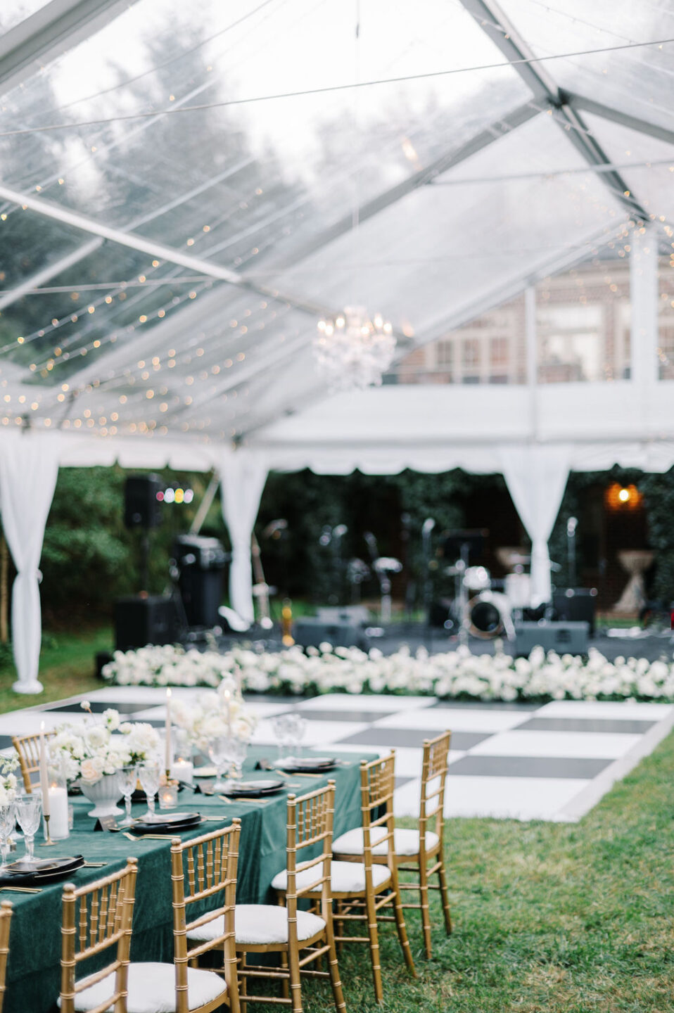 Clear-top wedding tent with bistro lights and a crystal chandelier, with a green-linen dinner table, gold chiavari chairs, a checkered dance floor and a band stage behind.
