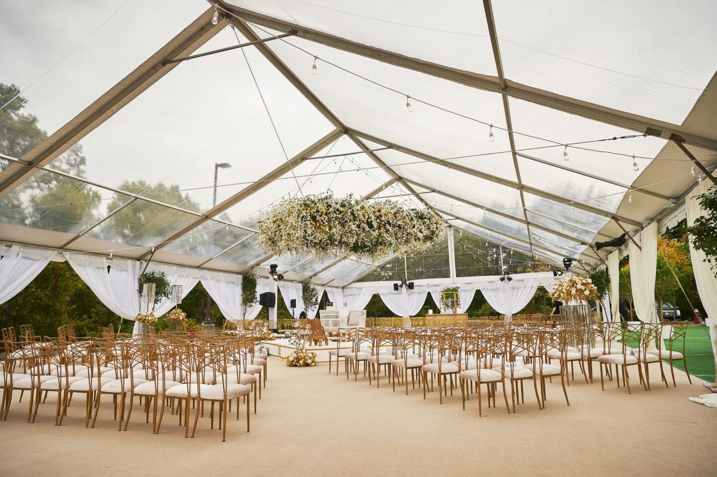Clear-top tent set for a wedding ceremony, with rows of gold chairs, a center aisle, a hanging floral installation and white draped sides. It's an example of event tent size for a ceremony layout.