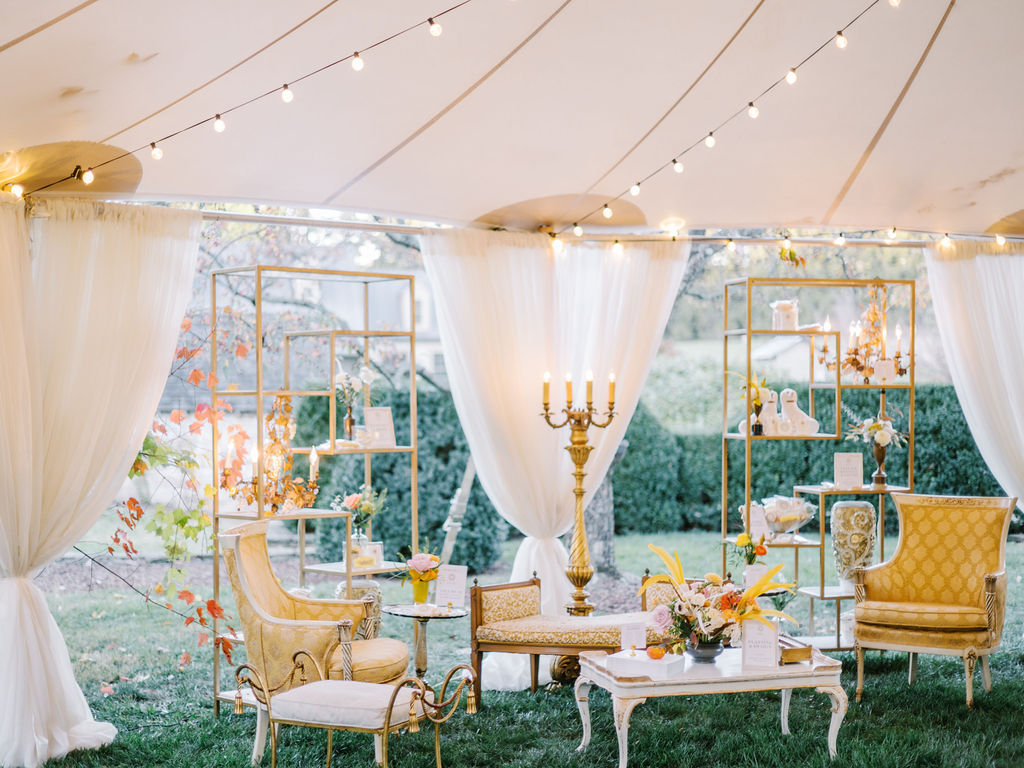 Lounge area under a sailcloth tent with gold shelving, vintage yellow armchairs and a settee, a candelabra, draped sides and string lights overhead.