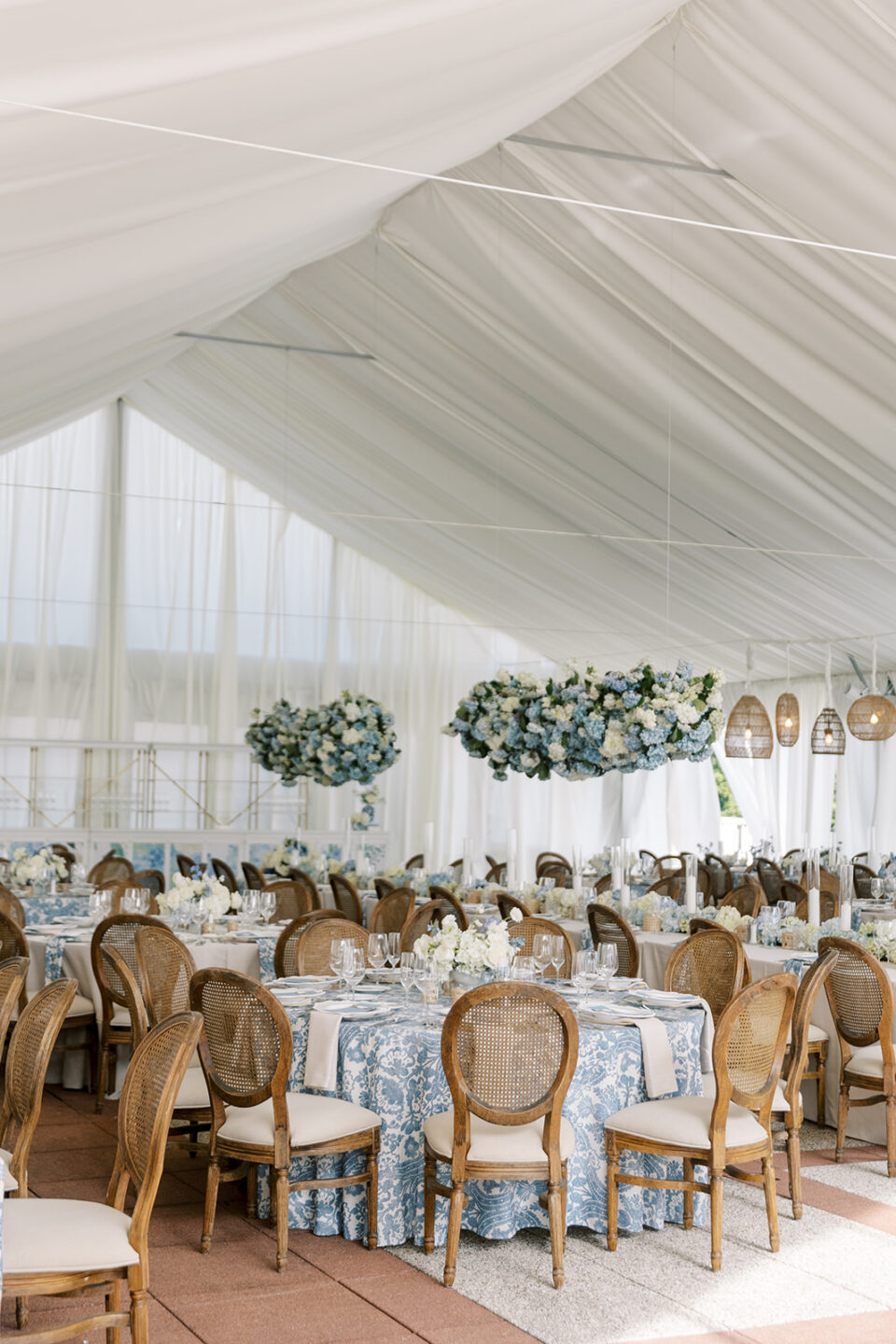 White draped tent set for a reception, with round tables in blue floral linens, cane-back chairs, hanging blue and white flower arrangements and rattan pendant lights. Use the event tent size chart to plan a seated dinner like this one.