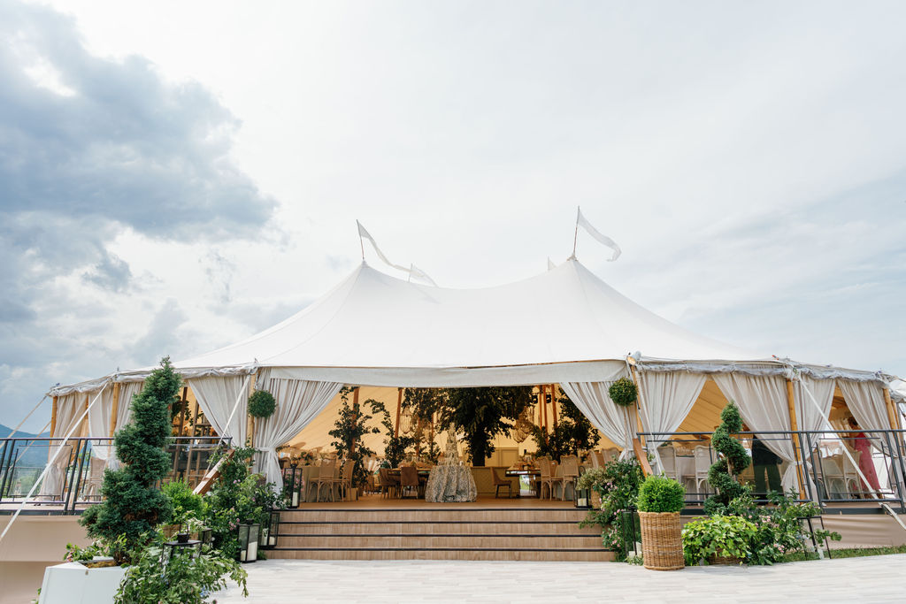 Outside view of a white sailcloth tent with flags on its peaks, open draped sides, entry steps and potted topiaries, with a dinner set up inside.