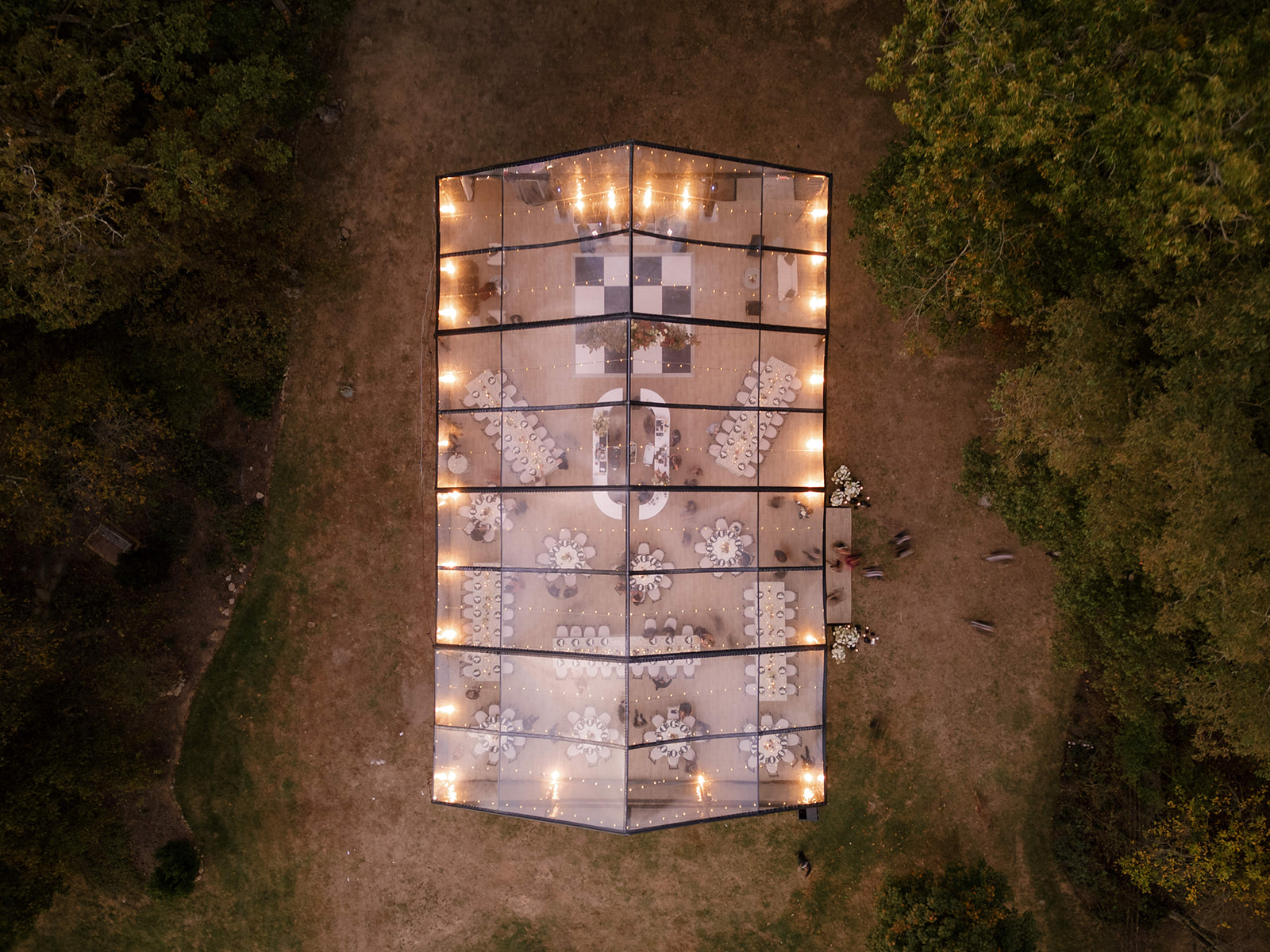Aerial view of a lit clear-top tent in a field, showing long and round dinner tables, a checkered dance floor and a lounge area. It shows how layout shapes event tent size.