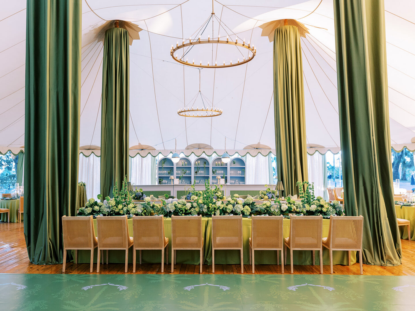 Sailcloth tent with green velvet draped poles, ring chandeliers and a long head table lined with cane chairs and greenery, next to a painted green dance floor.